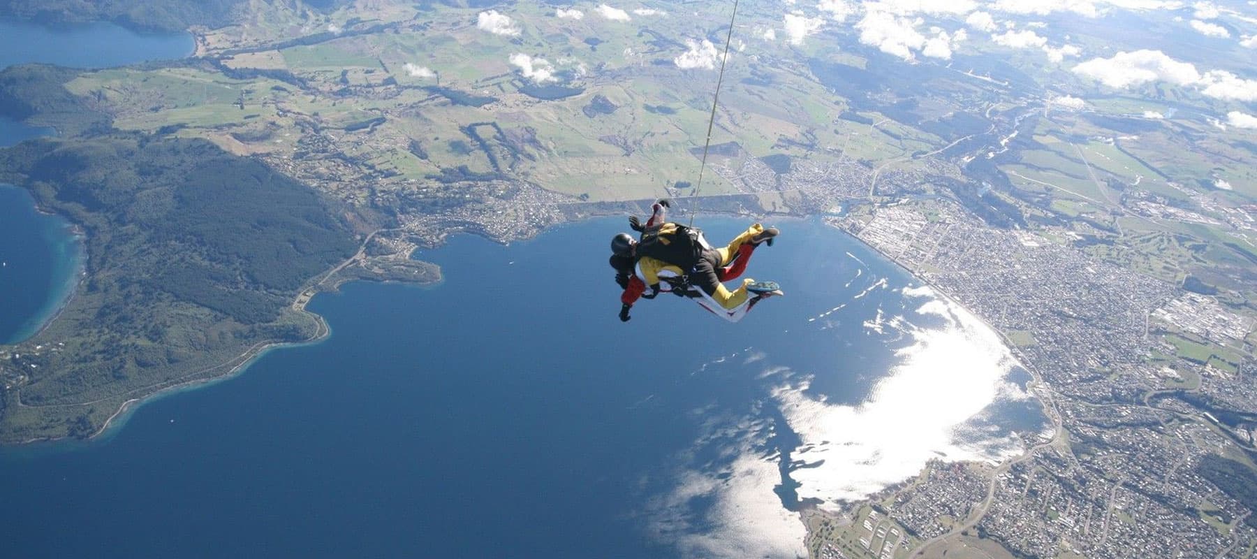 Two tandem skydivers descending above a large lake with surrounding greenery and hills, sunlight reflecting off the water below.