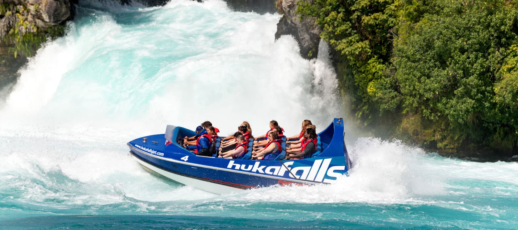  The image shows a blue jet boat labeled "hukafalls" speeding through turbulent rapids near a large waterfall. The boat is filled with passengers wearing red life jackets, gripping the seats as they navigate the powerful, churning water. The surrounding a
