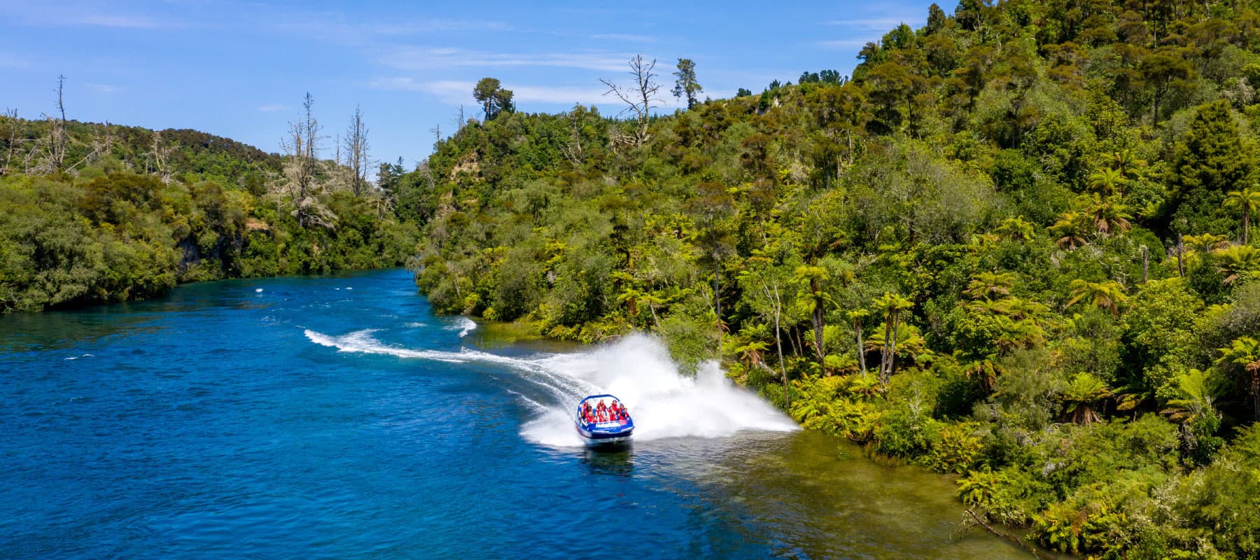  The image shows a blue jet boat labeled "hukafalls JET" speeding along a winding river, creating a large spray of water as it turns near the lush, green shoreline. The river is surrounded by dense vegetation, including tall trees and tropical plants, whi