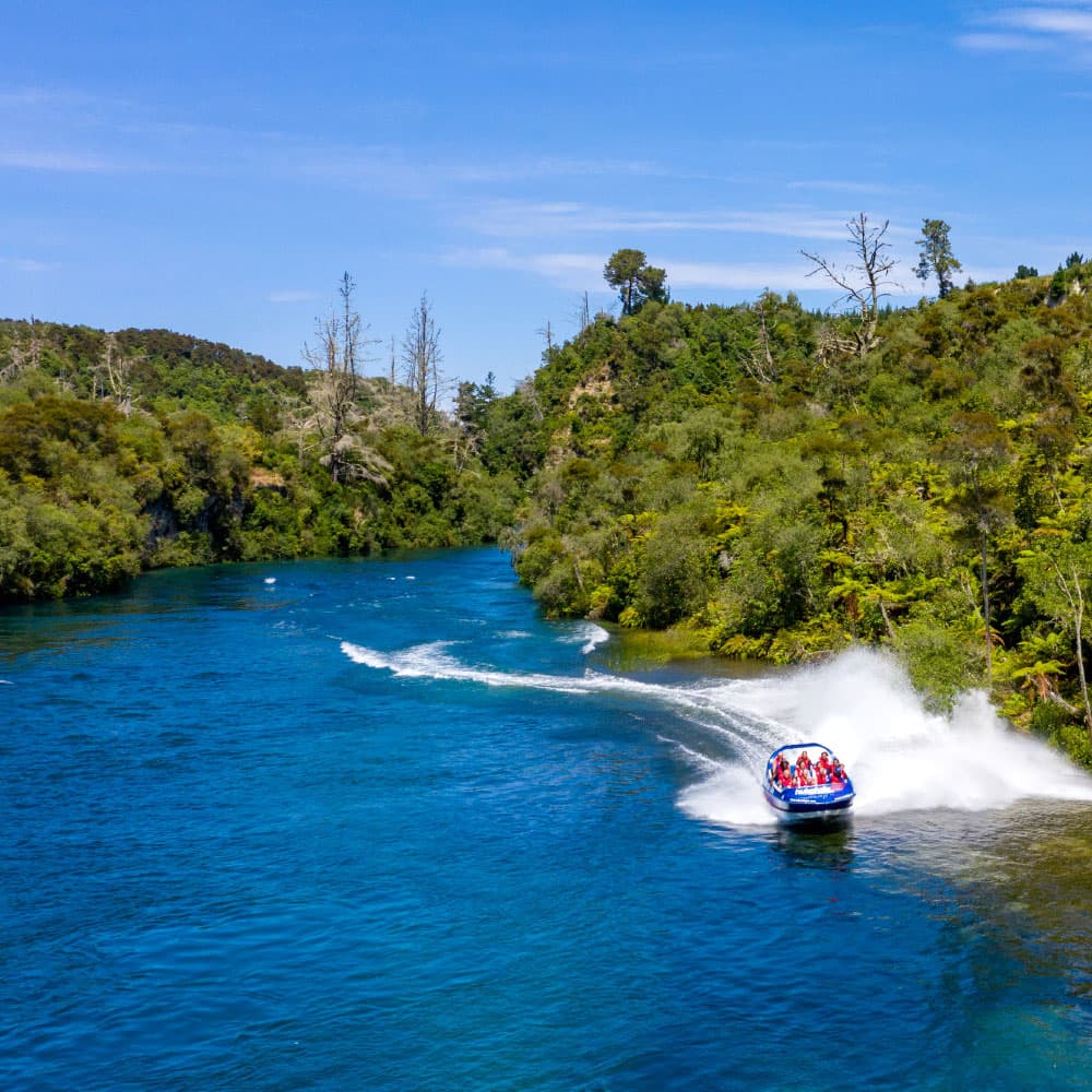 Jet boat racing on Waikato River near Huka Falls under clear blue skies, with turquoise water lined by native bush.