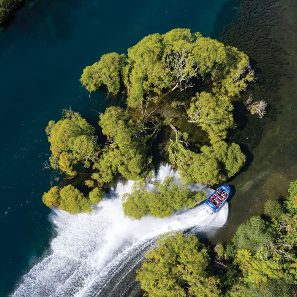 Aerial view of a Huka Falls Jet boat weaving through blue Waikato River surrounded by green trees in Taupō, New Zealand.